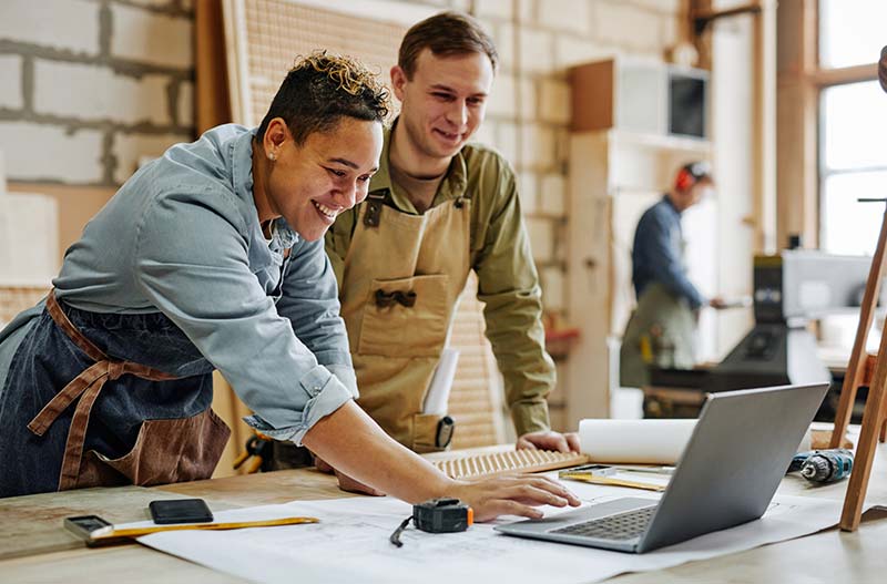 Warm toned portrait of animated carpenters using laptop and discussing furniture designs in workshop